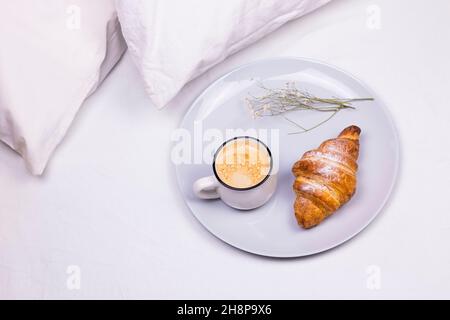 Tasse Kaffee mit Croissant und trockenen Blumen auf dem Teller. Morgens Frühstück Konzept im Bett Stockfoto