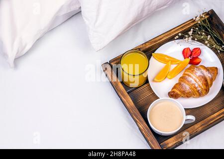 Kaffee, Orangensaft, Croissants, Milch, Erdbeere und Blumen auf dem Holztablett. Romantisches Frühstück am Morgen im Bett. Stockfoto
