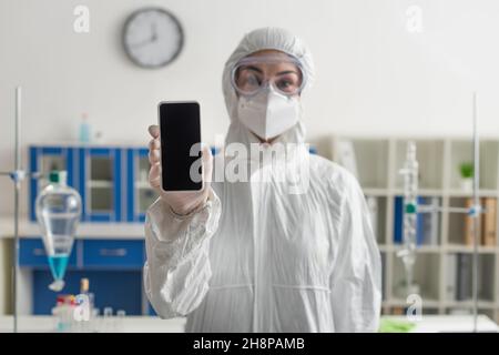 Verschwommener Arzt im Anzug mit Hazmat, der im Labor ein Mobiltelefon mit leerem Bildschirm hält Stockfoto