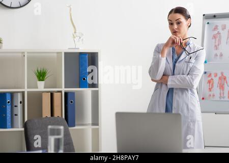 Nachdenklicher Arzt hält Brillen, während er in der Klinik auf einen verschwommenen Laptop schaut Stockfoto
