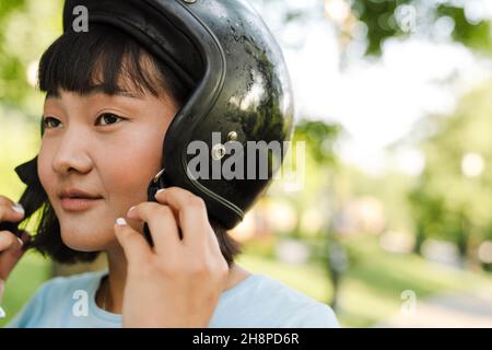 Nahaufnahme einer lächelnden jungen asiatischen Frau mit Motorradhelm im Park aus nächster Nähe Stockfoto