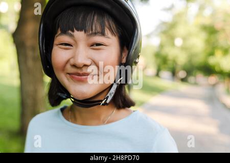 Nahaufnahme einer lächelnden jungen asiatischen Frau mit Motorradhelm im Park aus nächster Nähe Stockfoto