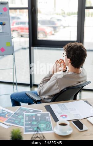 Nachdenklicher Geschäftsmann, der sich ein verschwommenes Flipchart in der Nähe von Dokumenten und Kaffee im Büro ansieht, Stockbild Stockfoto