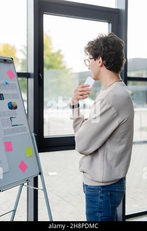 Seitenansicht eines jungen Geschäftsmannes, der sein Smartphone hält, während er im Büro auf das Flipchart schaut, Bild aus dem Stock Stockfoto