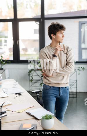 Nachdenklicher Geschäftsmann in einem Kopfhörer, der eine Brille in der Nähe des Arbeitstisches im Büro hält, Stockbild Stockfoto