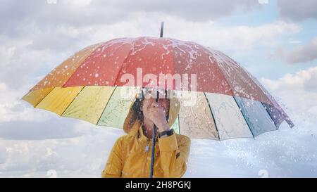 Glückliche junge Frau in gelbem Regenmantel unter Regenbogenschirm im spritzenden Wasser. Thema Meteorologie, Regen und schlechtes Wetter Stockfoto