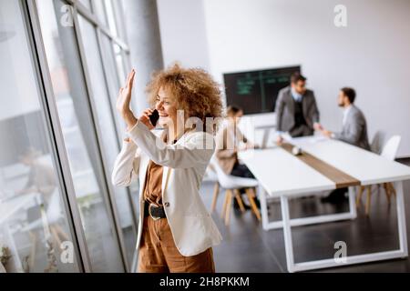 Junge Geschäftsfrau mit lockigen Haaren, die im Büro mit jungen Menschen ein Mobiltelefon benutzt, arbeitet hinter ihr Stockfoto