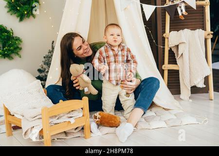 Junge, fürsorgliche und unterstützende Mutter, die mit einem aktiven, emotionalen Kind im Zelt im Zimmer spielt. Stockfoto