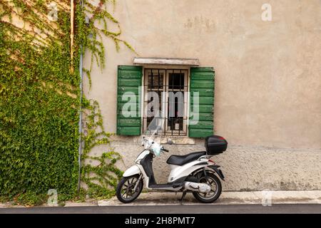 Typische italienische Straßenszene mit einem weißen Roller an einem Fenster mit grünen Fensterläden auf einer asphaltierten Straße in Colognola ai Colli, Verona, Italien. Stockfoto