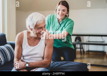 Glückliche Krankenschwester mit einem Stethoskop, das einen älteren Mann besucht, häusliche medizinische Assistentin Konzept, lächelnder Gesundheitsgast und ein älterer männlicher Patient, c Stockfoto