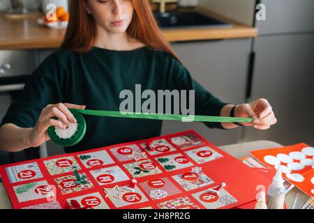 Ausgeschnittene Aufnahme einer fokussierten Rothaarige junge Frau, die zu Hause ein Weihnachtsband verwendet, um das Brett mit Adventskalender-Umschlägen zu umrahmen. Stockfoto