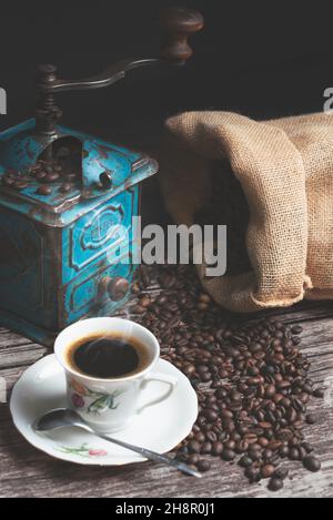 Vertikale Studioaufnahme des Stilllebens mit einer manuellen Kaffeemühle aus grünem Metall, Jute-Beutel und Kaffeebohnen auf einem alten Holztisch. Vintage weiße Tasse Stockfoto