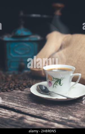 Vertikale Studioaufnahme des Stilllebens mit einer manuellen Kaffeemühle aus grünem Metall, Jute-Beutel und Kaffeebohnen auf einem alten Holztisch. Vintage weiße Tasse Stockfoto