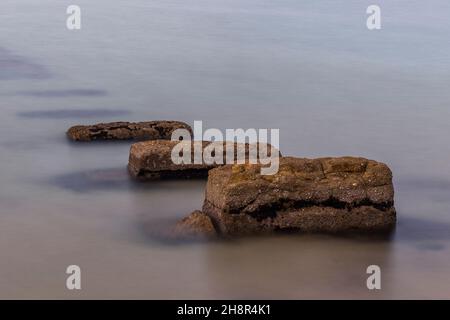 Große Felsen, die in einem milchigen oder glasartigen Meer untergetaucht sind, abstrakte große Felsbrocken im Meer, in der Größe abgestufte Felsen, die im Wasser verschwinden, Stockfoto