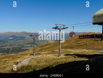 Die rote Gondel der Nevis Range-Seilbahn nähert sich an einem klaren, sonnigen Tag der Bergstation mit blauem Himmel, Hügeln und Wanderern im Hintergrund Stockfoto