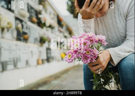 Trauernde Frau, die die Stirn berührt, während sie in der Nähe einer Säulengrube auf dem Friedhof sitzt Stockfoto