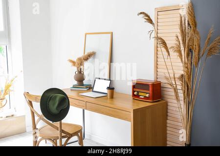 Moderner Arbeitsplatz mit Laptop und trockenem Schilf in Vase in der Nähe einer hellen Wand Stockfoto