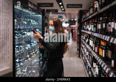 Weibliche Kundin in Maske liest Etikett auf Flasche Wein beim Einkaufen im Supermarkt während der Epidemie Stockfoto