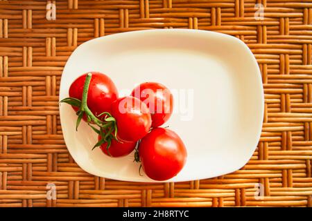 Eine Gruppe von roten Kirschtomaten auf einer weißen flachen rechteckigen Platte stehen auf einer hölzernen Korboberfläche. Blick von oben. Horizontales Foto. Lifestyle-Komposition. Nahaufnahme. Hochwertige Fotos Stockfoto