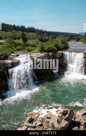 Porträtaufnahme des Wasserfalls von Lundbreck Falls mit Fluss, Brücke und blauem Himmel im Hintergrund Stockfoto