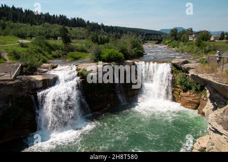 Landschaft aufgenommen Blick auf Lundbreck Doppel Wasserfälle mit Fluss, Brücke und kleinen Bergen im Hintergrund. Blauer Himmel grünen Bäumen Stockfoto