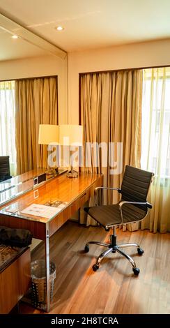 Ein moderner Arbeitsbereich mit einem Holztisch, einer Tischlampe, einem großen Spiegel und einem schwarzen Sessel in einem Hotelzimmer. Stockfoto
