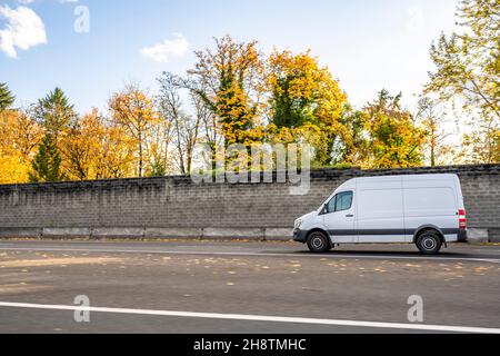 Kommerzielle kompakte Ladung kleiner weißer Mini-Van, der Ladung an den Kunden liefert, der auf der lokalen Straße mit herbstlichen gelben Bäumen und Betonschutzvorrichtungen unterwegs ist Stockfoto