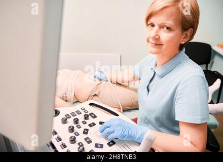 Gynäkologe in sterilen Handschuhen untersucht weibliche Patienten mit Ultraschallscanner, während Frau auf dem Tagesbett liegt. Ultraschalluntersuchung durch einen Ultraschalluntersuchung. Konzept der Ultraschalldiagnostik. Stockfoto