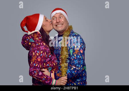 Liebevolles Paar mittleren Alters in festlichen Weihnachtsoutfits umarmen und küssen, während sie Weihnachten zusammen über einem grauen Backgrou feiern Stockfoto