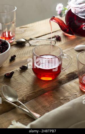 Mehrere transparente Gläser und Tassen sitzen auf einem Holztisch. Roter Tee aus Hibiskusblüten wird aus einer Glas-Teekanone in eine Tasse gegossen. Sudanesischer Rosenfl Stockfoto