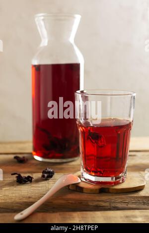 Auf einem Ständer auf einem Holztisch steht eine Glastasse mit kaltem Hibiskustee. Das Getränk hat dank der Blumen der sudanesischen Rose eine leuchtend rote Farbe. R Stockfoto