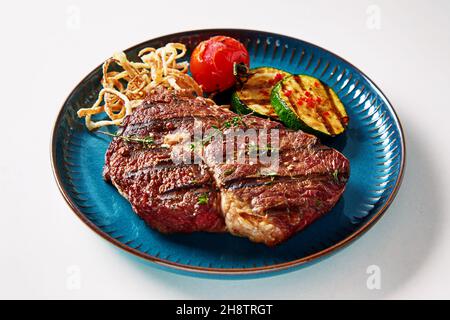 Auf blauem Teller auf weißem Hintergrund werden köstliche Grillgemüse und Rindersteak in einem hohen Winkel serviert Stockfoto