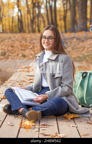 Junge schöne Schülerin sitzt in einem Herbstpark mit einem Lehrbuch Stockfoto