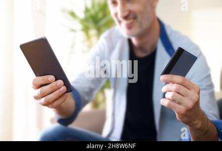 Konzept des Online-Shoppings mit Handy und Kreditkarte, wobei der Mann zu Hause mit Handy und Karte in der Hand sitzt. Horizontale Zusammensetzung. Stockfoto