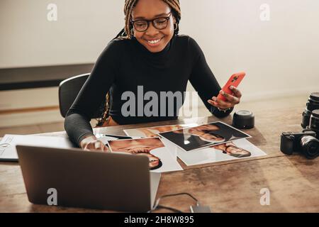 Künstlerin, die in ihrem Heimbüro einen Laptop und ein Smartphone verwendet. Kreative junge Fotografin, die fröhlich lächelt, während sie an ihrem Schreibtisch arbeitet. Freiberufliche Mitarbeiterin Stockfoto