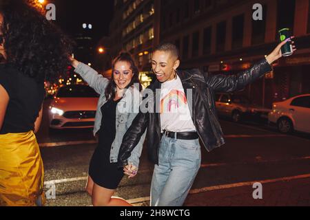 Tanz an einem Wochenendabend. Zwei fröhliche Freundinnen tanzen fröhlich, während sie eine Straße in der Stadt überqueren. Zwei lebhafte junge Frauen, die lachen und havi Stockfoto