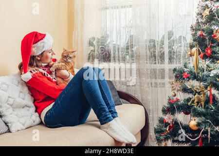 Christmas celebration with cat. Woman playing and hugging pet in Santa's hat by New year tree at home. Family relaxing during holiday Stockfoto