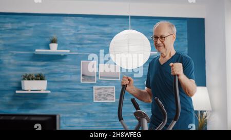 Ältere Person, die körperliche Übungen auf einem stationären Fahrrad macht, mit Fitnessgeräten trainiert. Älterer Mann, der zu Hause mit einem Cardio-Gerät die Beinmuskulatur trainiert und trainiert. Rentner Radfahren Stockfoto