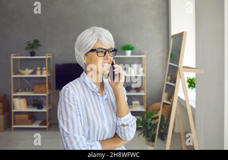 Glückliche ältere Frau, die im Wohnzimmer steht, Mobiltelefon hält, anruft und lächelt Stockfoto