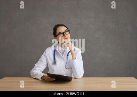 Porträt einer ernsthaften Ärztin, die mit einem nachdenklichen Gesichtsausdruck an einem Tisch sitzt. Stockfoto