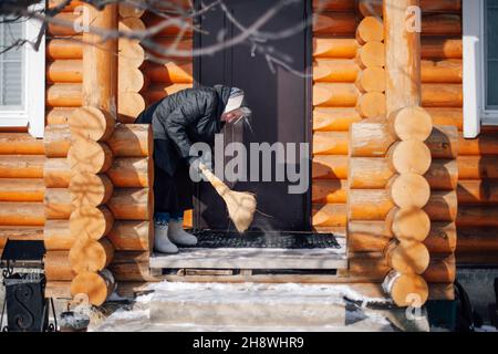 Seitenansicht der Frau auf der Veranda. Kaukasische Frau in Filzstiefeln und Schal hält Besen in ihren Händen und fegt im Hinterhof aus Holz heruntergefallenen Schnee Stockfoto