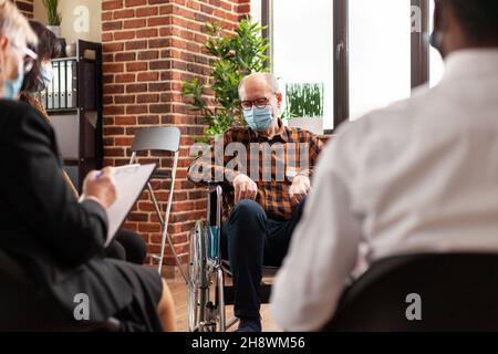Eine alte Person mit Gesichtsmaske sitzt im Rollstuhl beim Meeting-Programm. Mann mit Behinderung nimmt an einer Gruppentherapie Teil, um Alkoholabhängigkeit zu heilen und während einer Pandemie eine Beratung zu erhalten. Stockfoto