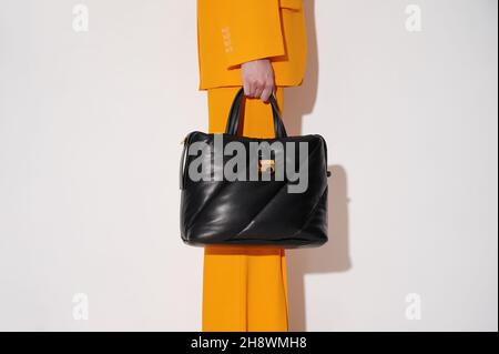 Frau in einem orangefarbenen Anzug mit schwarzer Ledertasche in der Hand auf weißem Hintergrund. Gesteppte Tragetasche modisches Accessoire. Neue Kollektion oder Verkauf. Stockfoto