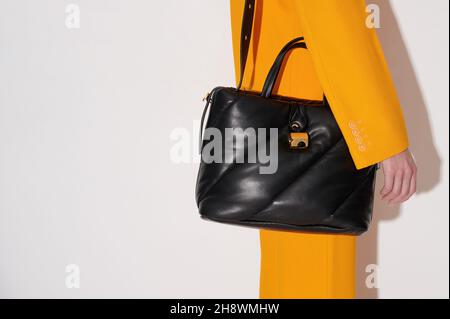 Frau in einem orangefarbenen Anzug mit schwarzer Ledertasche in der Hand auf weißem Hintergrund. Gesteppte Tragetasche modisches Accessoire. Neue Kollektion oder Verkauf. Stockfoto