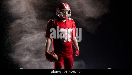 Amerikanischer Fußballspieler in roter Uniform hält Ball vor schwarzem Hintergrund mit Nebel Stockfoto