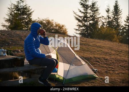 Seitenansicht eines Jungen in einer Kapuzenjacke, der Kaffee auf einer Holzbank in seinem Lager in der Nähe des Zeltes trinkt, das auf einer Bergwiese aufgestellt wurde. Herbstwandern und Camping in der Natur. Konzept von Wandern, Reisen und Camping. Stockfoto