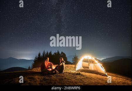 Frau mit Gitarre in den Händen, die am Lagerfeuer neben ihrem beleuchteten Zelt gegenüber dem Mann sitzt. Romantischer Abend im Freien unter Sternenhimmel in den Bergen. Konzept des Nachtcampens und Beziehungen. Stockfoto