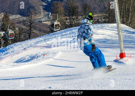 Ein Mann rutscht auf einem Snowboard eine Skipiste hinunter. Schnee fliegt unter seinem Brett heraus. Vor der Kulisse der Seilbahn. Wintersport während der vac Stockfoto