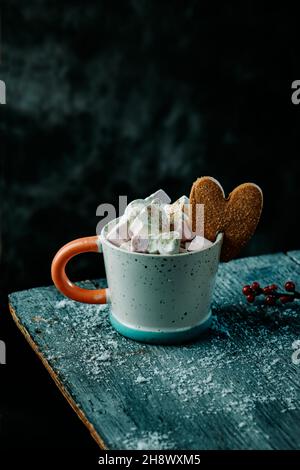 Eine Tasse Kaffee, gekrönt mit einigen Marschwalben und einem herzförmigen Lebkuchenkekse, neben einem Zweig winterberry auf einem grauen rustikalen Holztisch spr Stockfoto