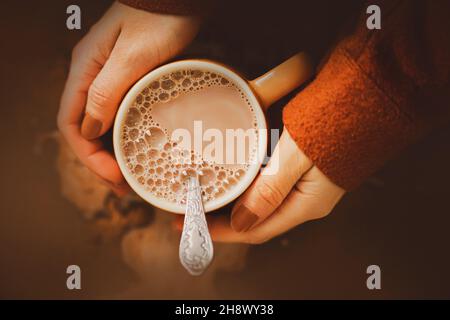 Eine Frau in einer braunen Wolljacke hält einen Becher warmen duftenden köstlichen Kakaos mit Milch in der Hand. Ein angenehmer Zeitvertreib beim Frühstück. Ein warmes Getränk in kalten wir Stockfoto